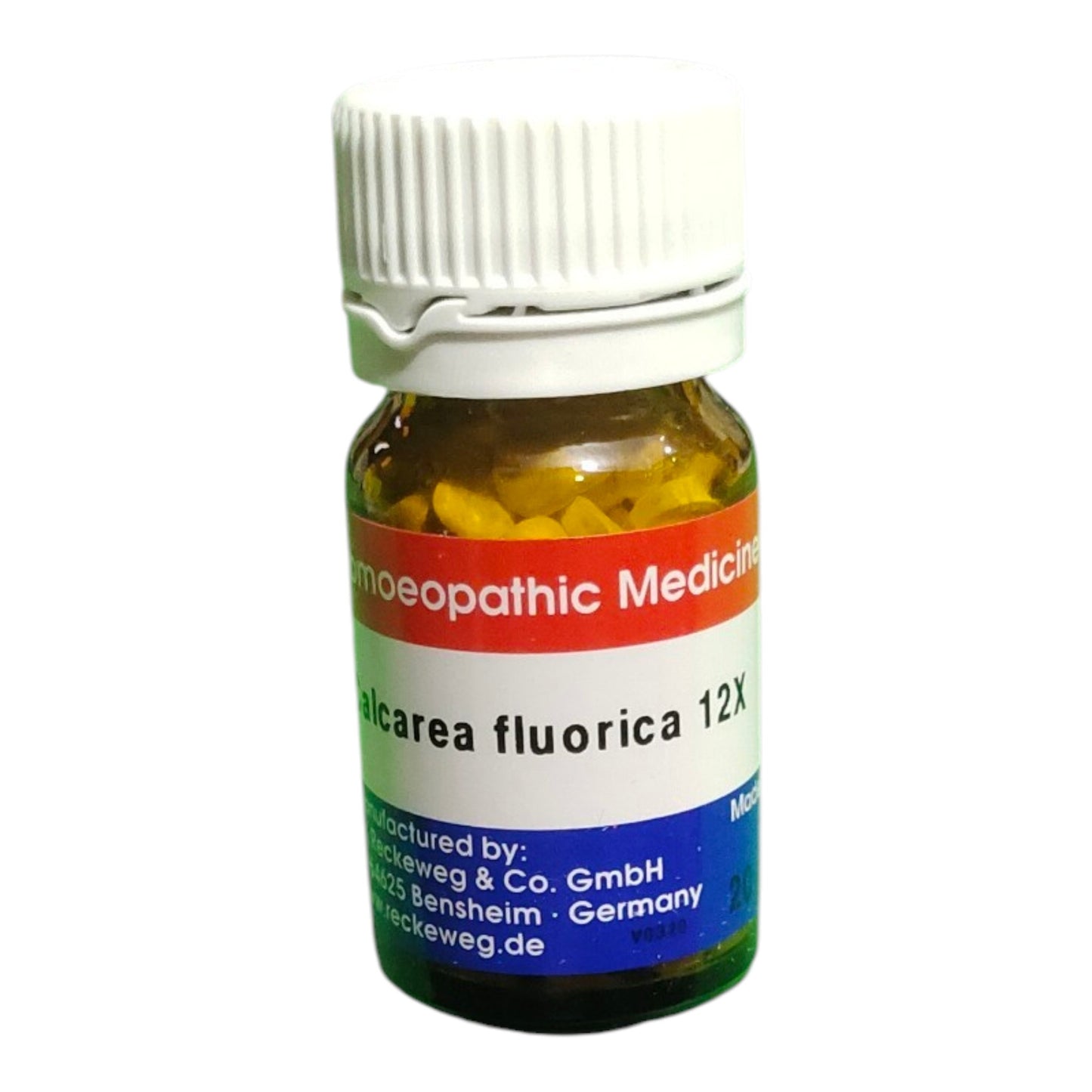 A small amber glass bottle of homeopathic medicine with a white ribbed cap, showing yellow tablets inside. The label features red, white, and blue sections with German text, including 'homeopathic Medicin' and 'alcarea fluorica 12X'.